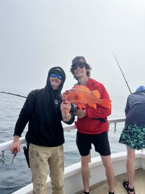 Two anglers proudly displaying a vibrant Rockfish while fishing on a boat in foggy conditions.