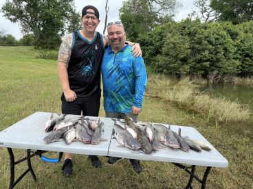 Two anglers proudly posing with a catch of multiple catfish displayed on a table after a successful fishing trip.