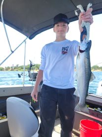 A young angler proudly displaying a freshly caught trout while fishing on a boat, surrounded by a scenic water backdrop.
