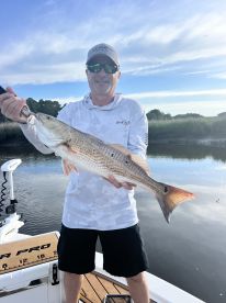Angler proudly displaying a large Redfish while standing on a boat in a serene waterway.