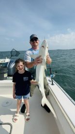 A proud angler and a young girl display a large shark caught during a fishing trip on a boat, showcasing a family fishing experience.