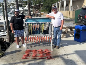 Two anglers proudly displaying a variety of fish, including Snapper and Mahi Mahi, on a catch board at the marina after a successful fishing trip.