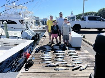 A group of young anglers proudly posing with their catch of various fish displayed on the dock after a successful fishing trip.