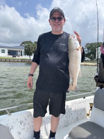 An angler proudly displaying a Redfish while on a fishing trip, with a scenic waterfront in the background.