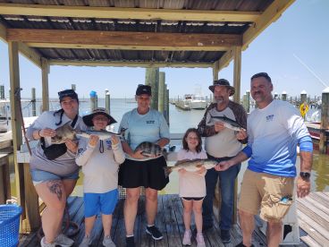 A family group proudly displaying their catch of various fish, including a notable Speckled Trout, at the dock after a successful fishing trip.