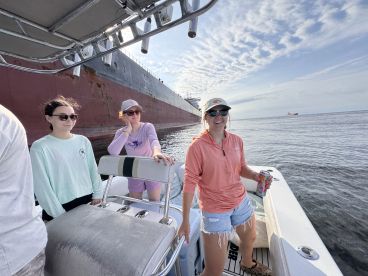 Group of young women enjoying a day on the water, with a large cargo ship in the background, showcasing a relaxed fishing atmosphere.
