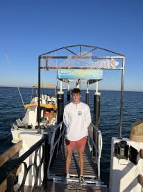 A young man stands on the dock in front of a fishing boat, showcasing the vibrant 'Vive el Sueño' sign against a clear blue sky.