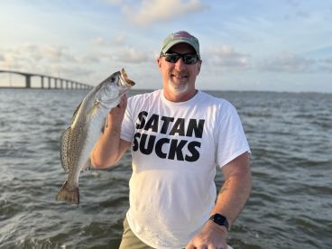 An angler proudly displaying a large Speckled Trout while fishing near a bridge, showcasing a beautiful sunset backdrop.