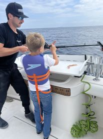 A young boy in a life jacket is learning to fish with guidance from an adult on a boat, showcasing a hands-on fishing experience.