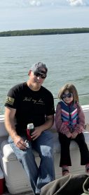 A father and daughter enjoying a day on the water, seated comfortably inside a boat, with a scenic backdrop of greenery and water.