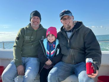 A family enjoying a fishing trip on a boat, with a young girl in a life jacket smiling between two adults.