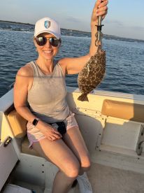 A smiling angler holding a Flounder while sitting on a boat, enjoying a sunny day on the water.