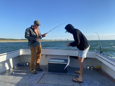 Two anglers actively fishing on a boat, with one reeling in a catch while the other prepares to assist. The scene captures the excitement of a fishing trip in a coastal environment.