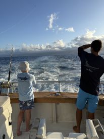Two young anglers enjoying a fishing trip on a boat, casting lines into the open ocean under a clear sky.