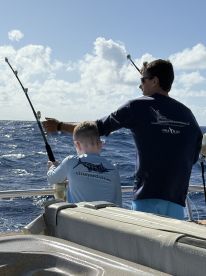A young boy and an adult are engaged in fishing on a boat, with the ocean and clouds in the background, showcasing a family fishing experience.