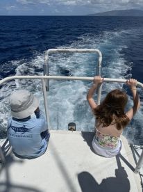 Two young anglers enjoying the boat ride, seated at the back of a fishing vessel, with the ocean in view and fishing gear visible.