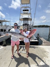 Two anglers proudly displaying their catch, including a large Kingfish and several Snapper, at the dock after a successful fishing trip.