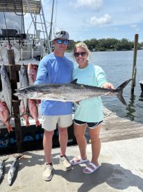 Two anglers proudly displaying a large King Mackerel alongside a variety of other fish at the dock after a successful fishing trip.