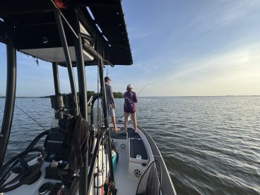 Two young anglers actively fishing from a boat in calm waters, showcasing a beautiful sunset backdrop.