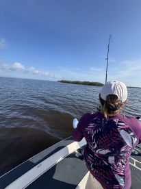 An angler casting a line from a boat in calm waters, surrounded by lush greenery in the distance.