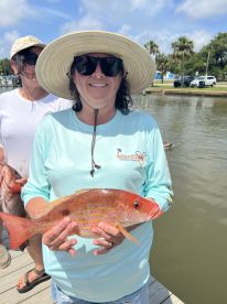 An angler proudly displaying a vibrant Red Snapper while standing on the dock, with another person in the background, showcasing a successful fishing trip.