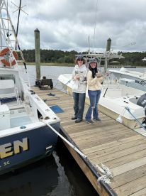 Two anglers proudly displaying their catch on the dock, with a fishing boat in the background, showcasing a successful day on the water.
