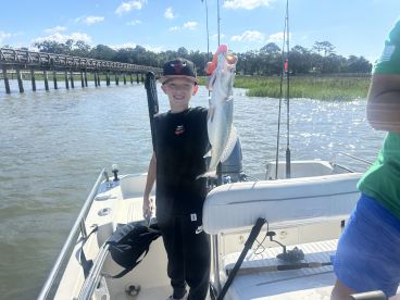 A young angler proudly displaying a caught Speckled Trout while fishing from a boat near a scenic bridge.