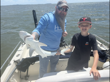A young angler and an adult proudly displaying a caught shark while on a fishing boat, enjoying a sunny day on the water.