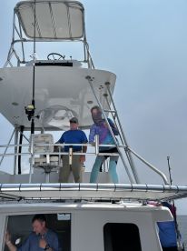Two anglers on the upper deck of a sportfishing boat, preparing for a day on the water. The boat features a spacious helm area and multiple fishing rods ready for use.