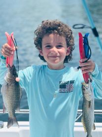Young angler proudly displaying a large striped bass and a speckled trout while fishing on a boat.