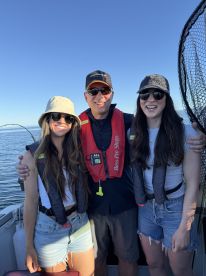 Three anglers enjoying a sunny day on the water, wearing life jackets and smiling aboard a fishing boat.