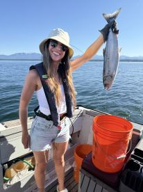 Angler proudly displaying a large salmon while fishing on a sunny day, with scenic mountains in the background.