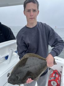 A young angler proudly displaying a large Flounder while fishing on a boat, showcasing a successful catch in overcast weather.