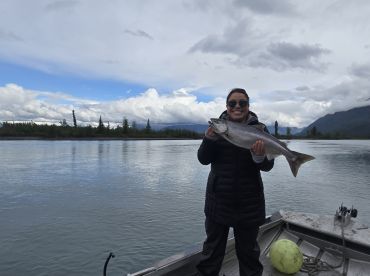 A happy angler holding a large salmon while fishing on a scenic river, surrounded by lush greenery and mountains.