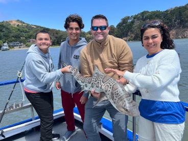 Family enjoying a successful fishing trip, proudly displaying a Leopard Shark on a boat in a scenic coastal environment.