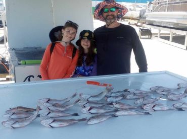A family poses with their catch of multiple fish on a boat's cleaning table, showcasing a successful fishing trip.