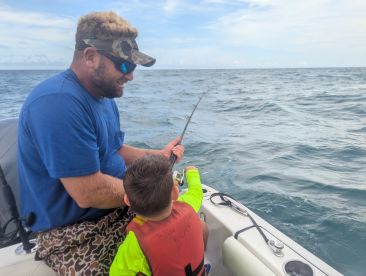 A young angler and an adult are engaged in reeling in a fish while fishing on a boat in open waters, showcasing a fun and educational fishing experience.