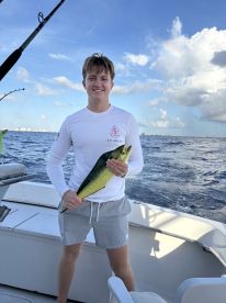 A young angler proudly displaying a vibrant Mahi Mahi aboard a fishing boat in clear blue waters.
