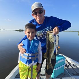 A young boy in a life jacket proudly poses with his grandfather, showcasing a large Largemouth Bass caught during a serene day on the water.