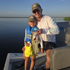 A young angler and an adult proudly displaying a large Largemouth Bass while fishing on a calm lake.