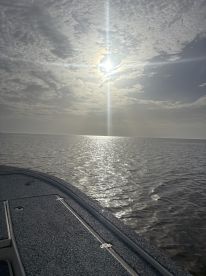 A serene view of the boat's side against a reflective water surface, with a cloudy sky and sun peeking through, ideal for a peaceful fishing day.