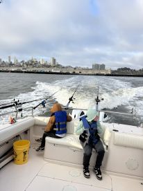 Two young anglers seated inside a boat, wearing life jackets, as they prepare for a fishing adventure in a scenic urban harbor.