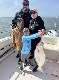 A family of four proudly displaying a large Flounder on a fishing trip, with the boat's interior visible in the background.