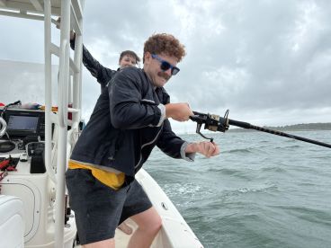 Two anglers engaged in fishing action on a boat, with one reeling in a catch while the other supports from behind. The scene captures the excitement of fishing in a cloudy environment.