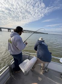 Two anglers engaged in a fishing battle on a boat, with a bridge visible in the background, showcasing a dynamic fishing environment.