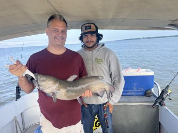 Two anglers proudly displaying a large Catfish while fishing on a boat in a calm waterway.