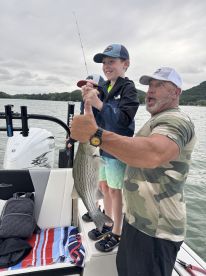 A young angler joyfully holds a striped bass with the help of an adult on a fishing boat, showcasing a successful catch during a family outing.