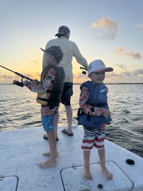 Two young boys fishing with an adult on a boat during sunset, showcasing a family fishing experience.