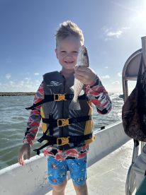 A young angler proudly displaying a caught fish while enjoying a sunny day on the water, wearing a life jacket and colorful swim shorts.