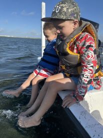 Two young boys enjoying a day on the water, sitting on the edge of a boat with their feet in the water, showcasing a fun fishing experience.